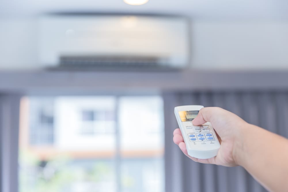 Main ajustant la température de la climatisation avec une télécommande vers un système de climatisation mural pour rafraîchir la maison.